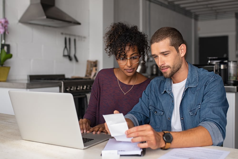 young couple sits at kitchen table looking at a laptop and receipts