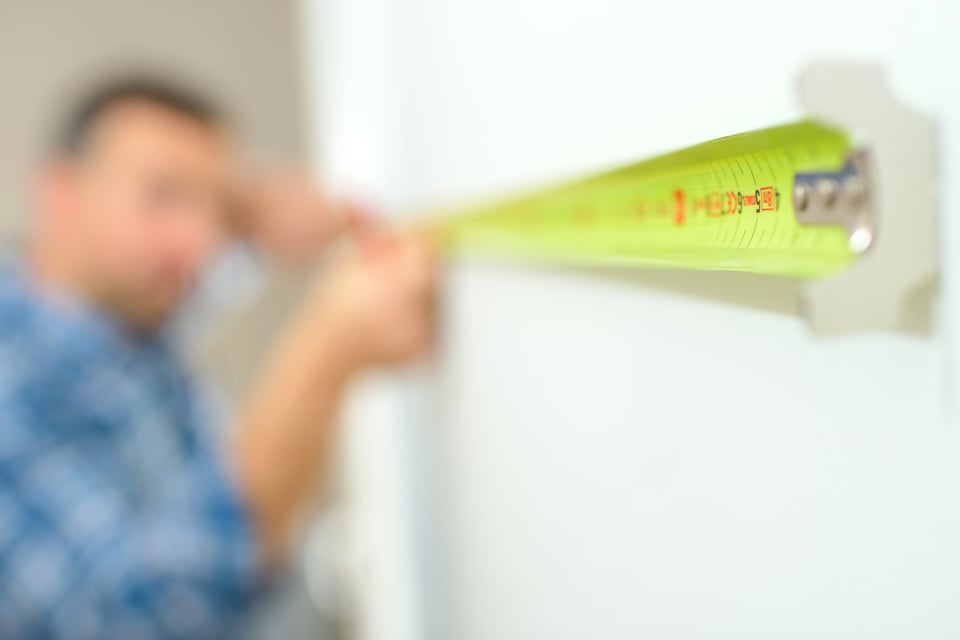 close up on the end of a retractable measuring tape with a man blurry in the distance eyeing up a measurement on a wall