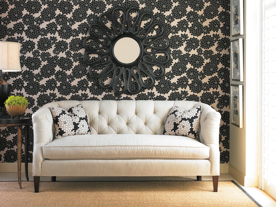 neutral sofa with tufted back and floral accent pillows with a side table and a plant in front of busy floral wall paper and an accent mirror on the wall