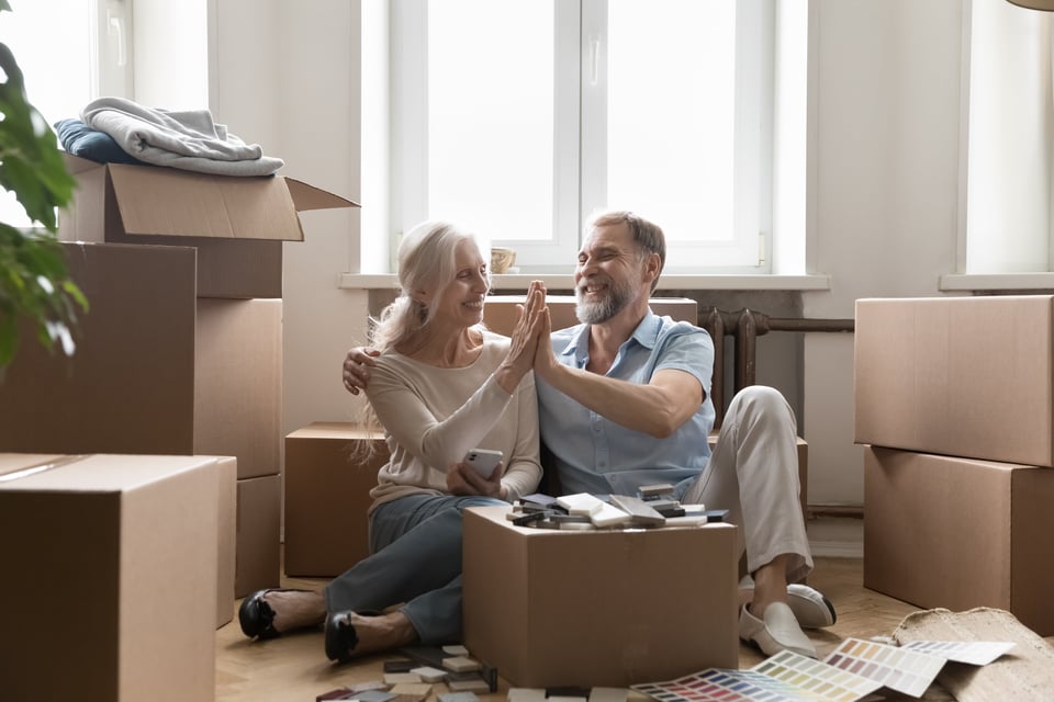 Senior couple of home owners discussing interior design improvement, clapping hands, giving high five over moving box, decoration samples, hugging, laughing, sitting on color in new apartment
