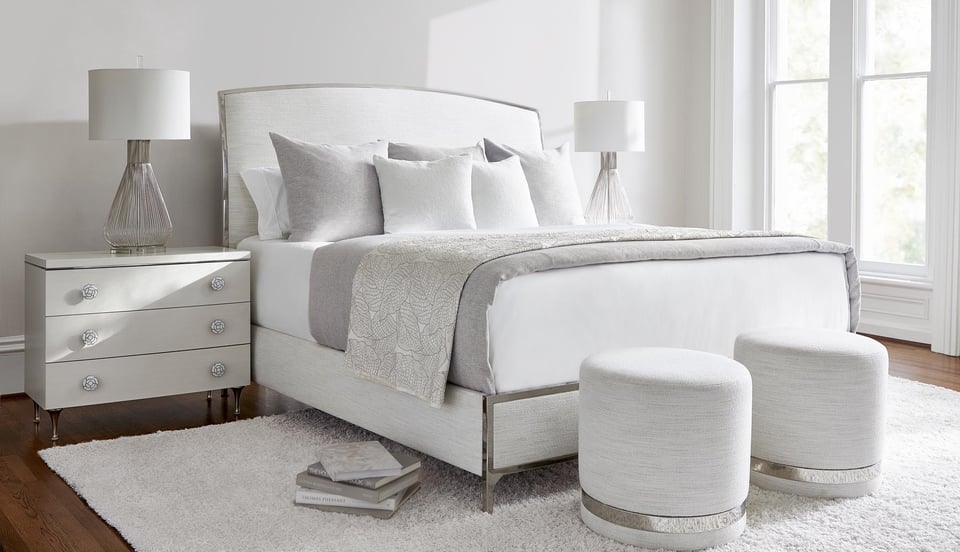 bedroom suite with upholstered headboard, footboard, and sideboard, two small ottomans at foot of the bed over a shag area rug and and two nightstands with matching lamps