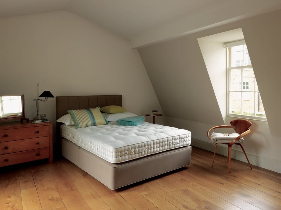 bedroom with a bed and an undressed mattress over top of a divan foundation, pillows and a folded blanket over top with a dresser, chair, pedestal table and lamp around the bed