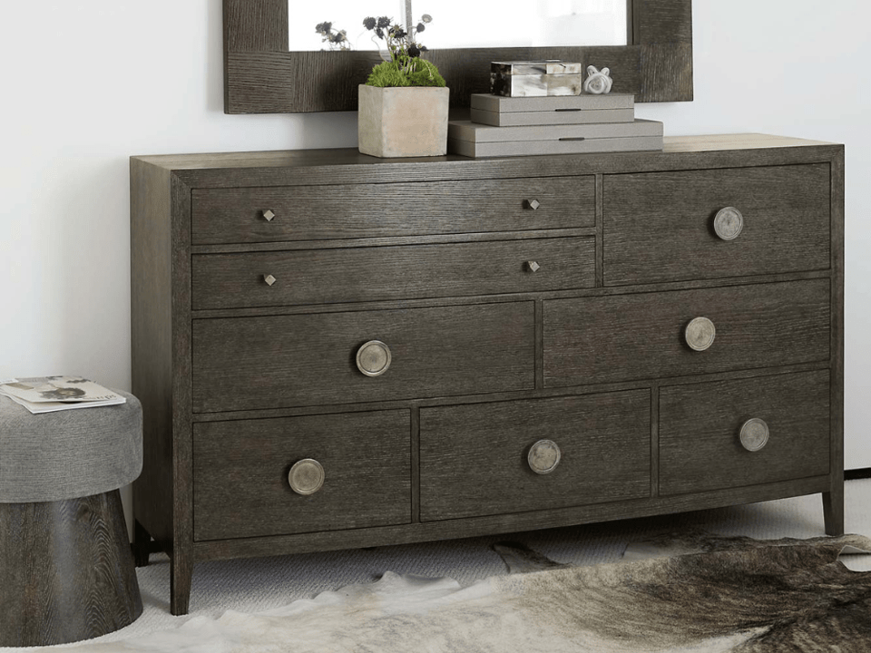 wood dresser with eight drawers of various sizes in front of a mirror with a nearby upholstered stool