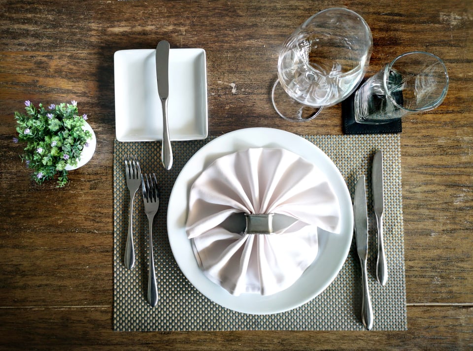 place setting with place mat, two forks, three knives, wine glass, drinking glass with coaster, small plate, and a decorative plant over a wood table