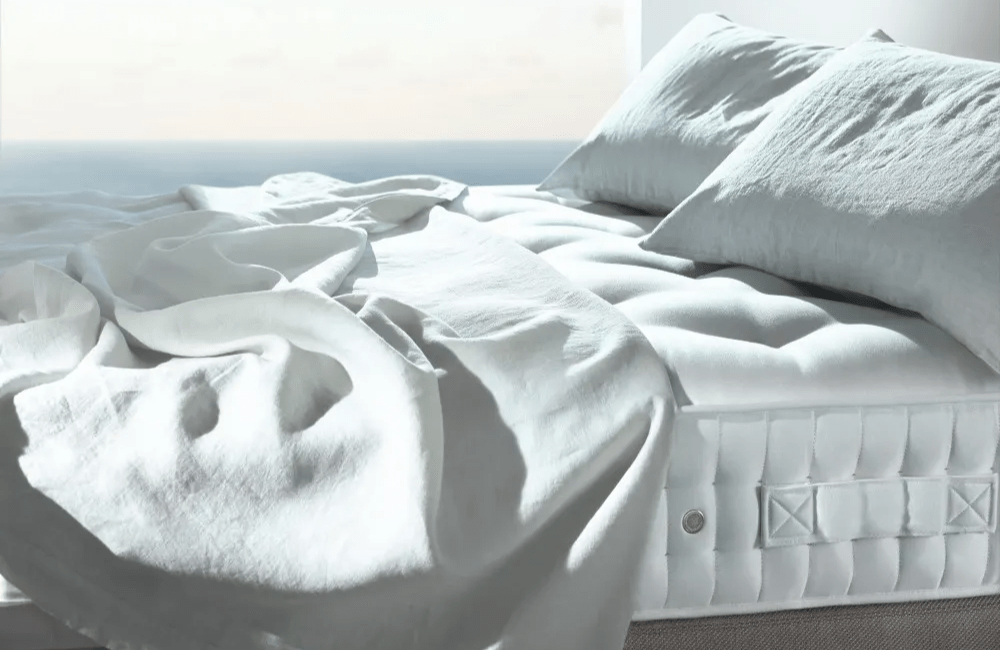 mattress with a loose sheet on top and two pillows in front of an oceanfront window