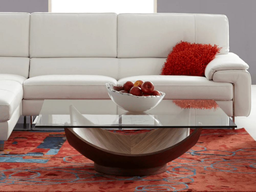 table with glass top and concaved wood base, bowl filled with apples on top over an area rug with a sofa and throw pillow behind